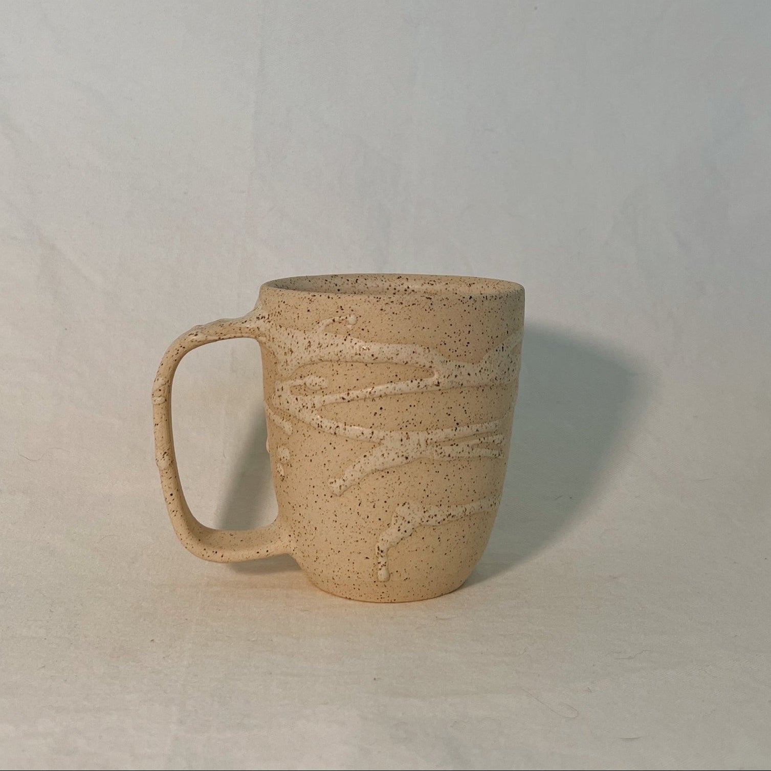 Off-white ceramic mug with handle, with white glaze drizzle, on a plain white background